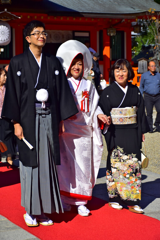 Жених в черной накидке хаори с гербами семьи и штанах - хакама в узкую полоску. Невеста - в сиромуку – кимоно белого цвета, которое является символом чистоты и непорочности девушки. Головной убор - ватабоси. Рядом мама невесты.