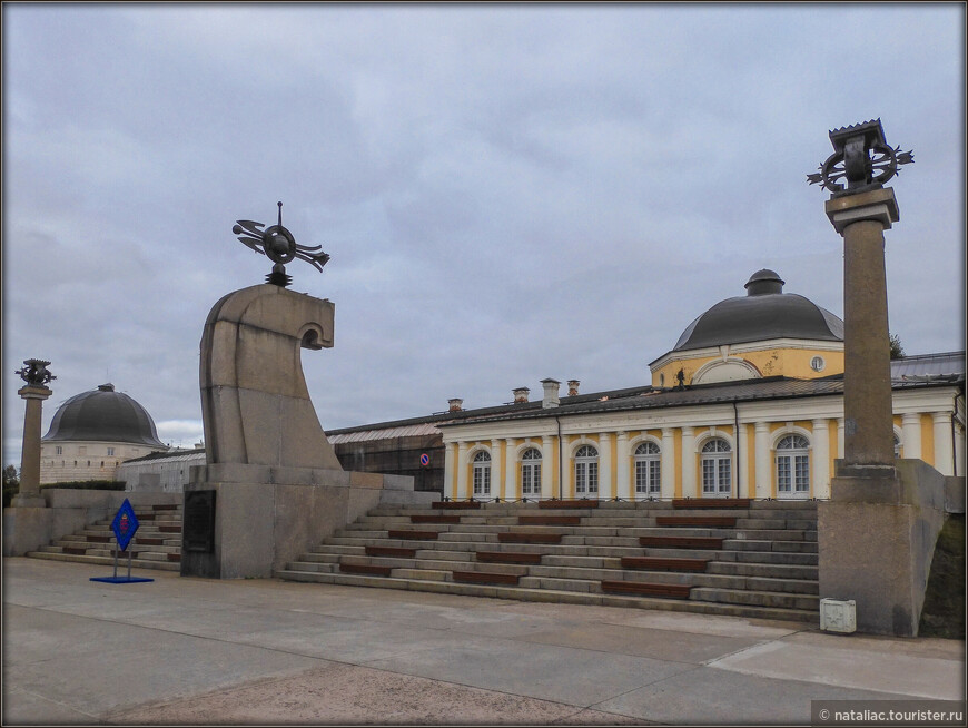 Памятник, напоминаюющий морскую волну, установленный на месте крепости, заложенной в 1584 году по приказу царя Ивана IV Грозного, рядом с которой и начал впоследствии расстраиваться город Архангельск.  На заднем плане остатки Архангельского гостиного двора построенного в1668—1684 годах. 