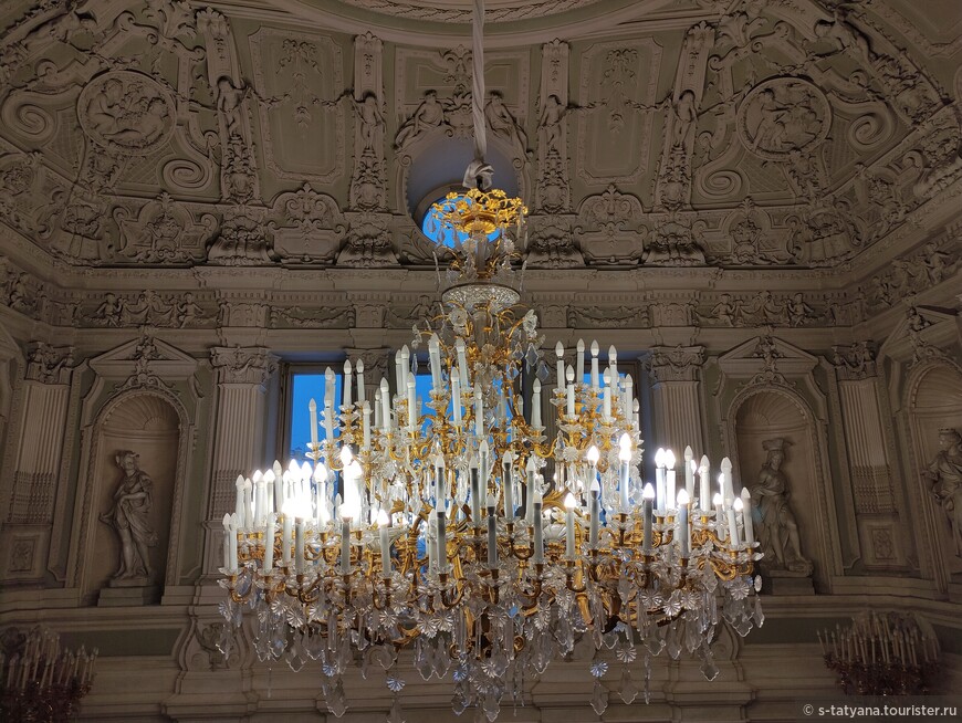 Освещает нижний вестибюль огромная люстра на цепях в стиле 18 века из позолоченной бронзы с плоской чашей гранёного стекла.